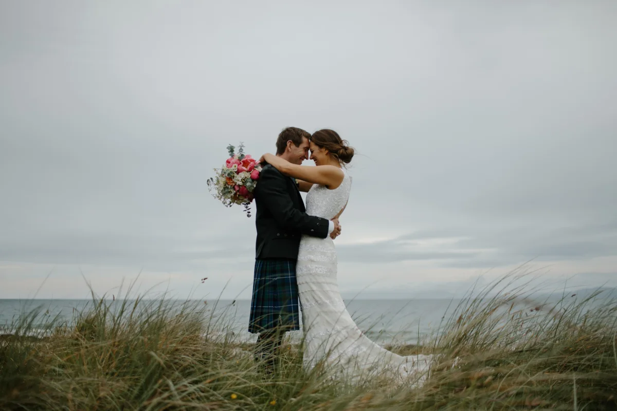 A couple embraces among dune grasses by the sea, the bride holding a bouquet.