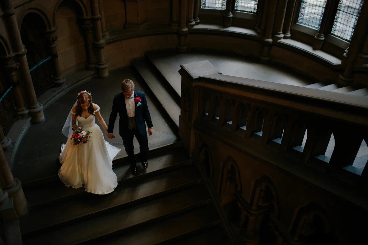Seen from above, a bride and groom walk down a dark wooden staircase.