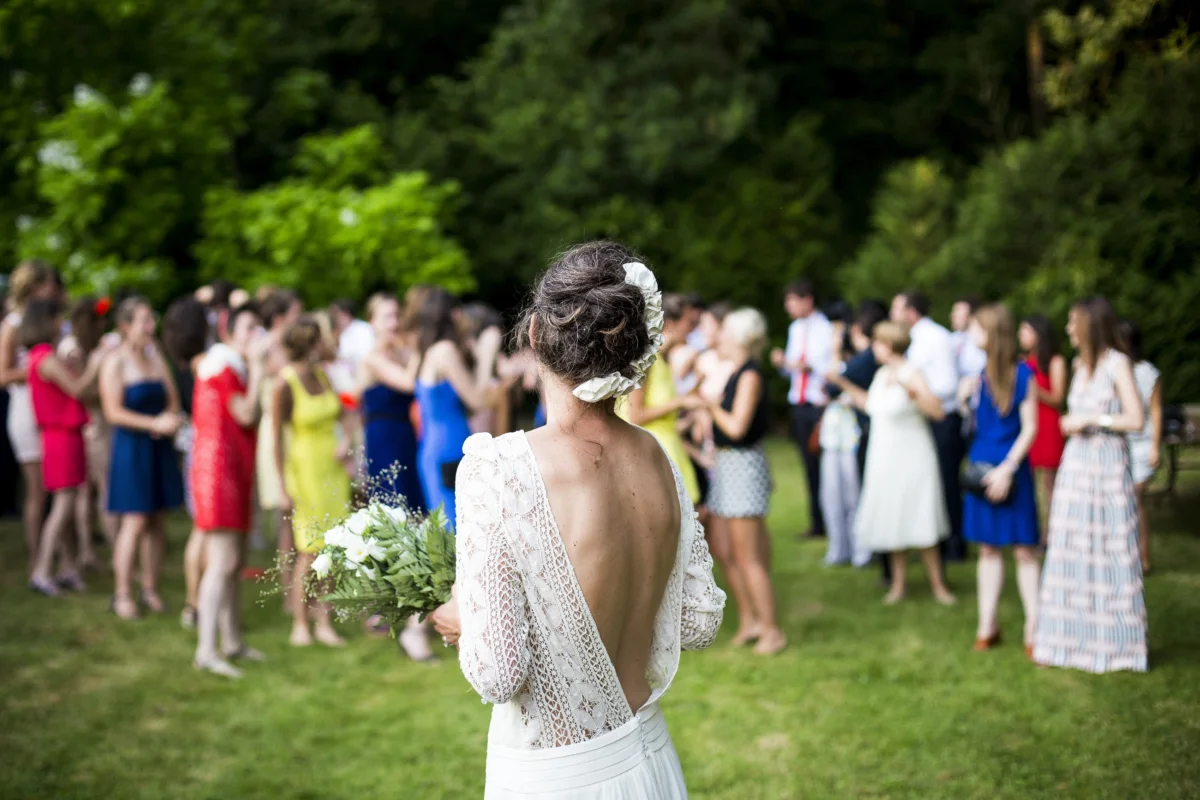 A bride seen from behind faces her guests on a garden lawn, bouquet in hand.