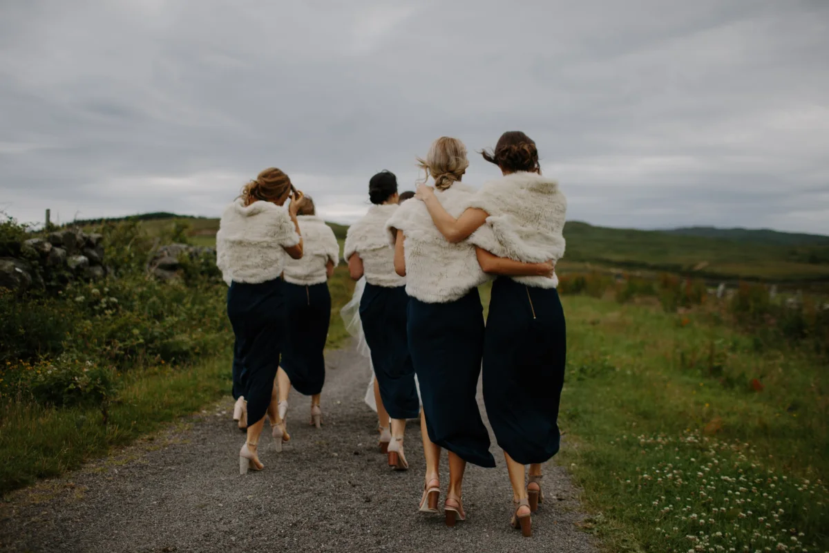 Four bridesmaids walk arm in arm down a country path.