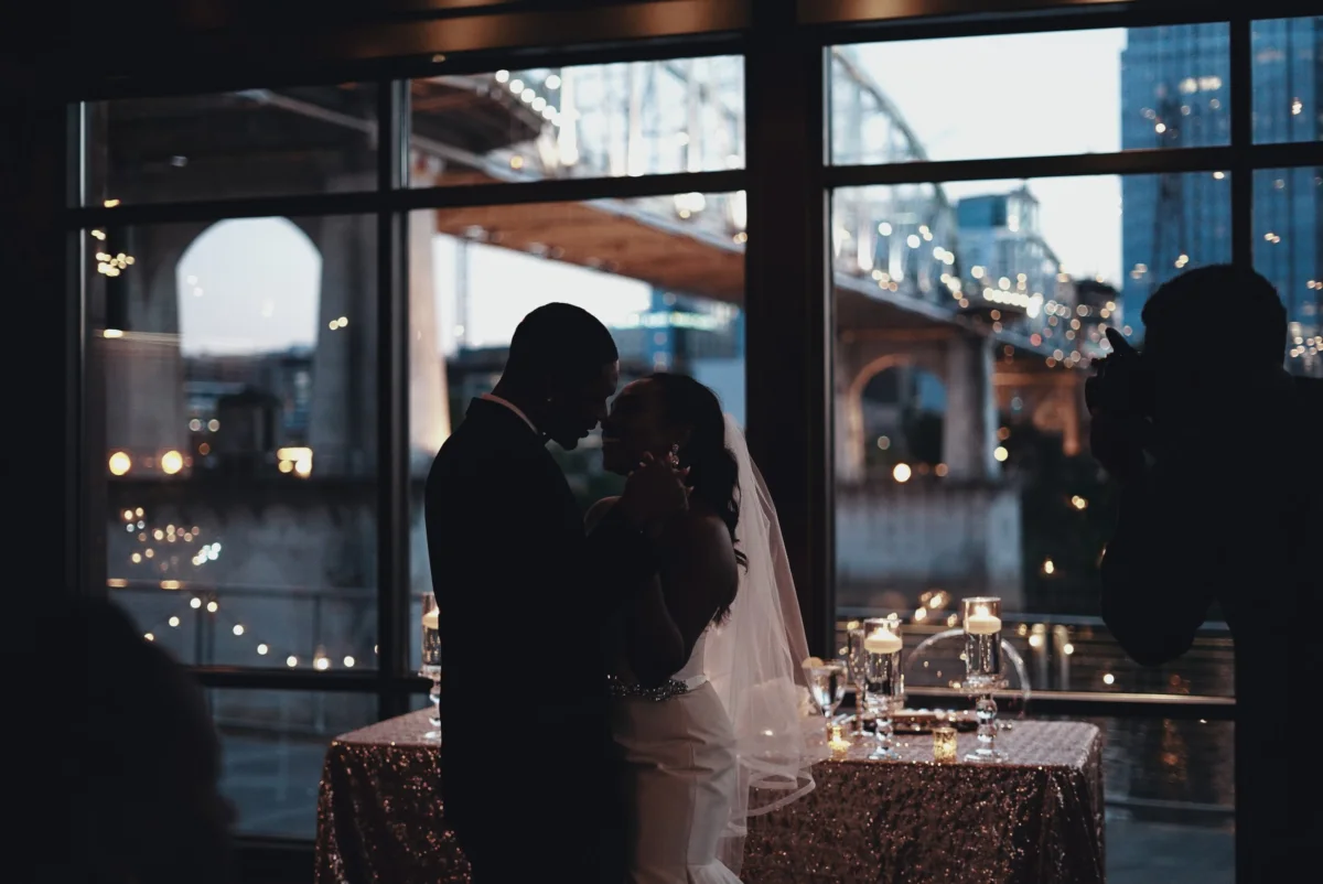 A couple in silhouette holds each other by a tall window in the evening.