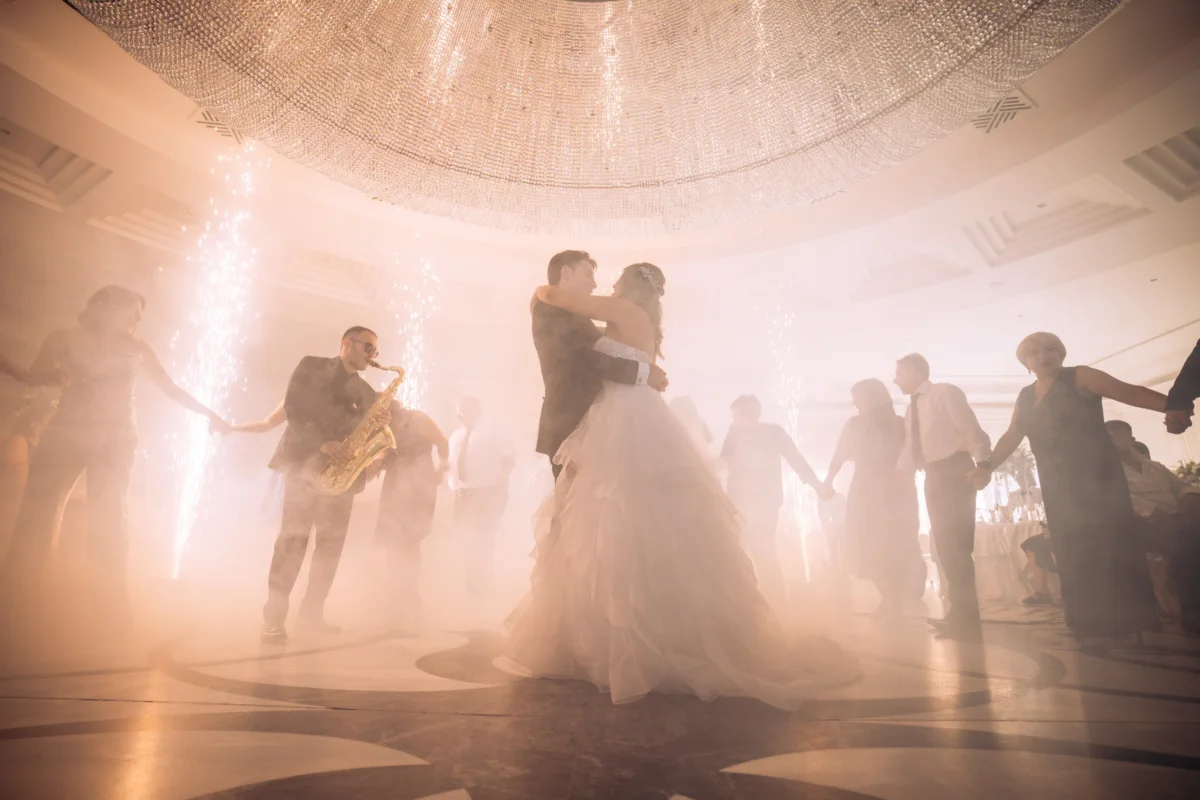 A couple shares their first dance in soft haze, guests and a saxophonist around them.