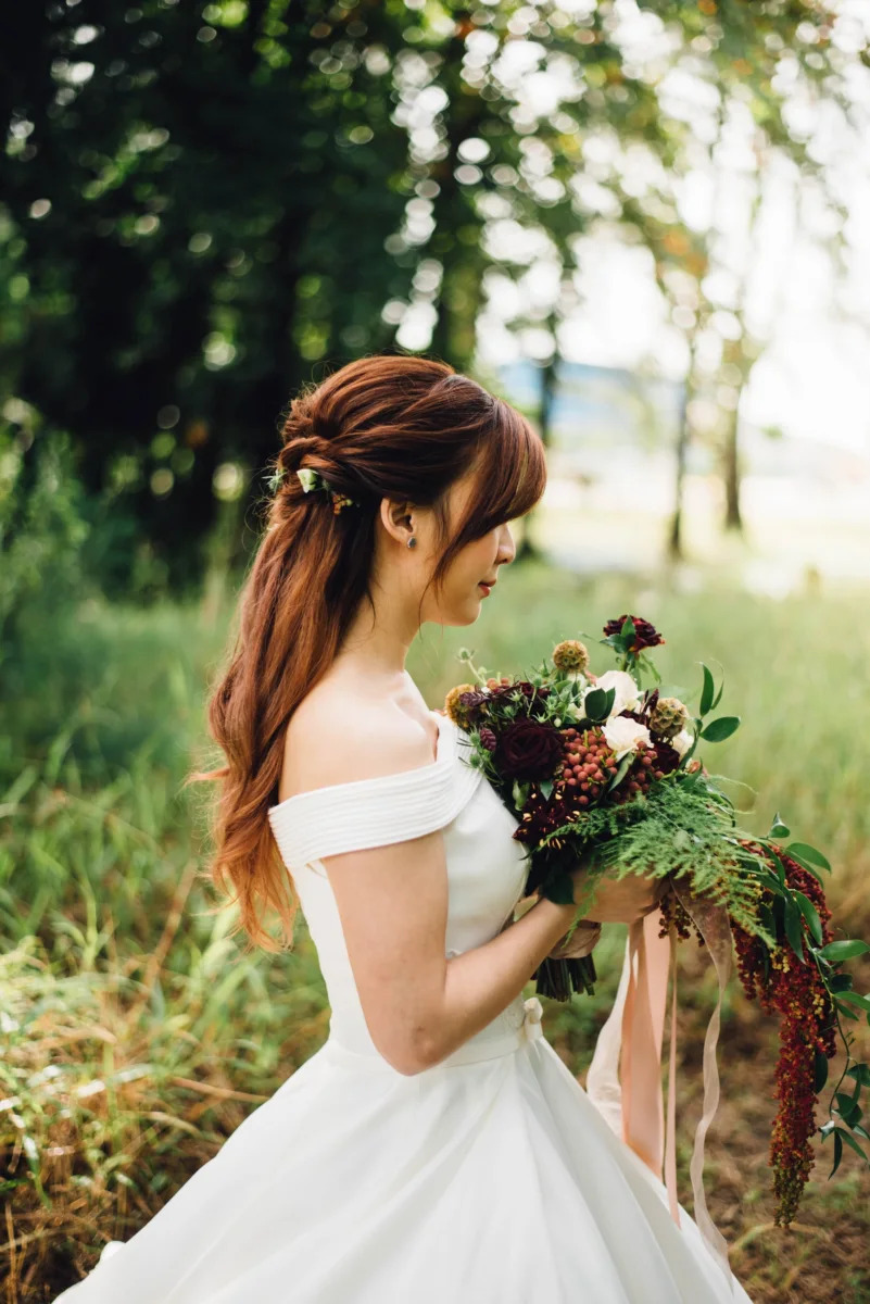 A bride in an off-shoulder gown looks down at her bouquet in a sunlit meadow.