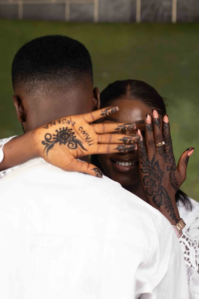 A bride covers her eyes with hennaed hands, smiling, as the groom holds her close.