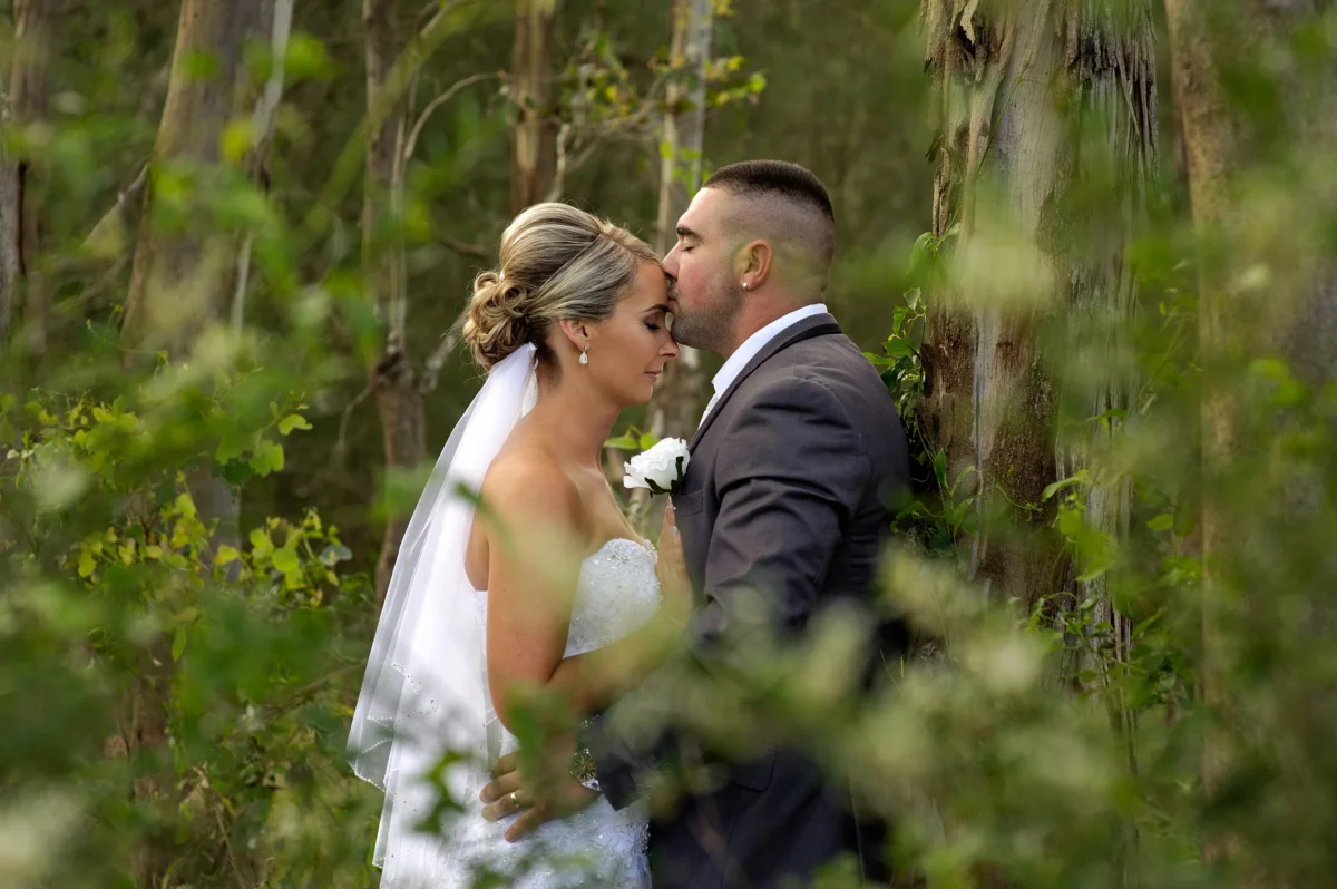 A groom kisses the bride's forehead among the trees.