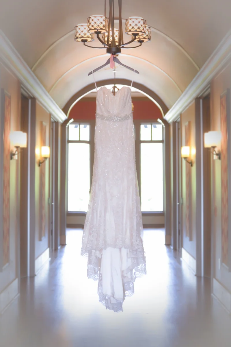 A lace wedding gown hangs from a chandelier in a bright, arched hallway.
