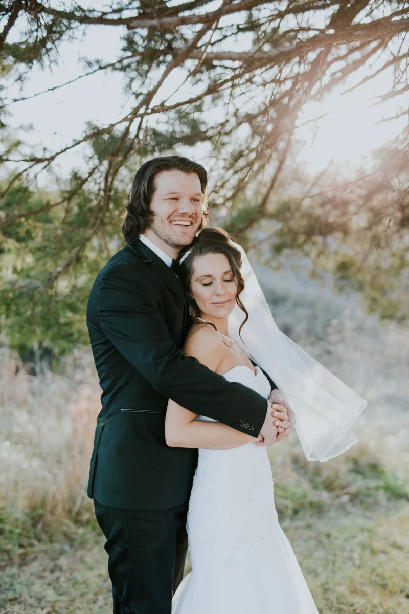 A groom wraps his arms around the bride from behind; she leans back with her eyes closed.