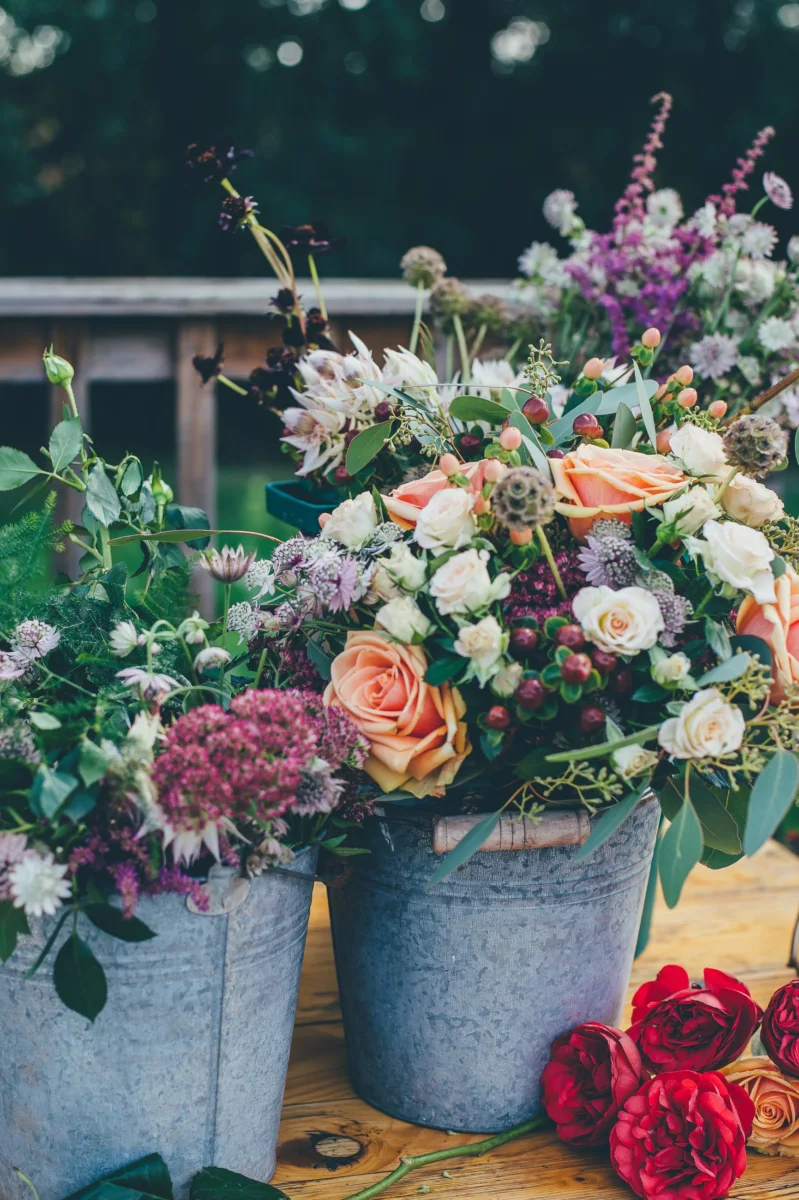 Garden roses and wildflowers arranged in galvanised buckets on a wooden table.