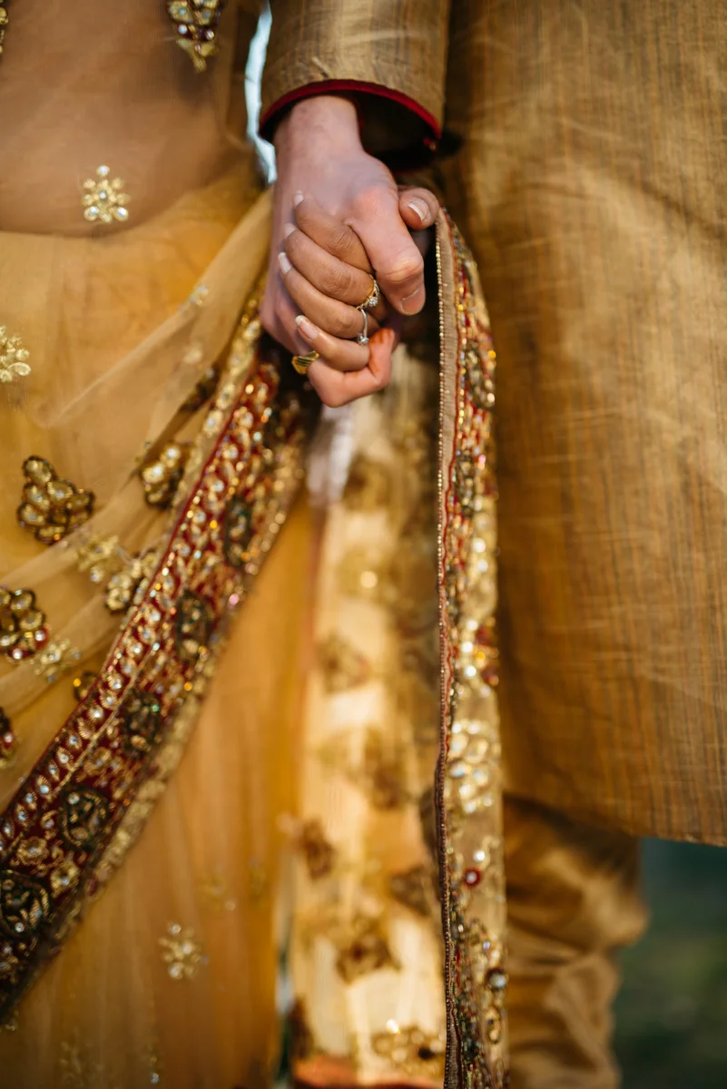 A couple in embroidered gold wedding clothes holds hands, seen up close.