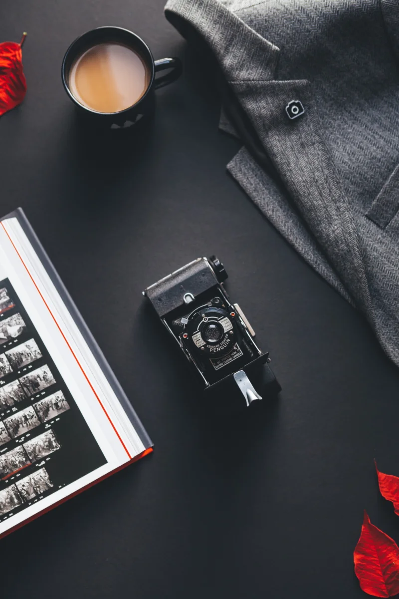 A vintage folding camera on a dark table beside a contact sheet and a cup of coffee.