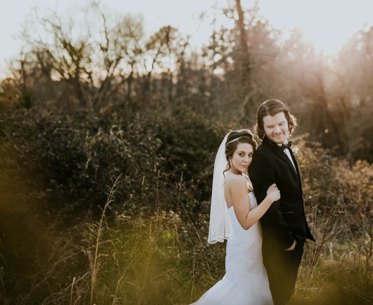 A bride in a veil leans against her groom's shoulder in tall grass, lit by low golden sun.